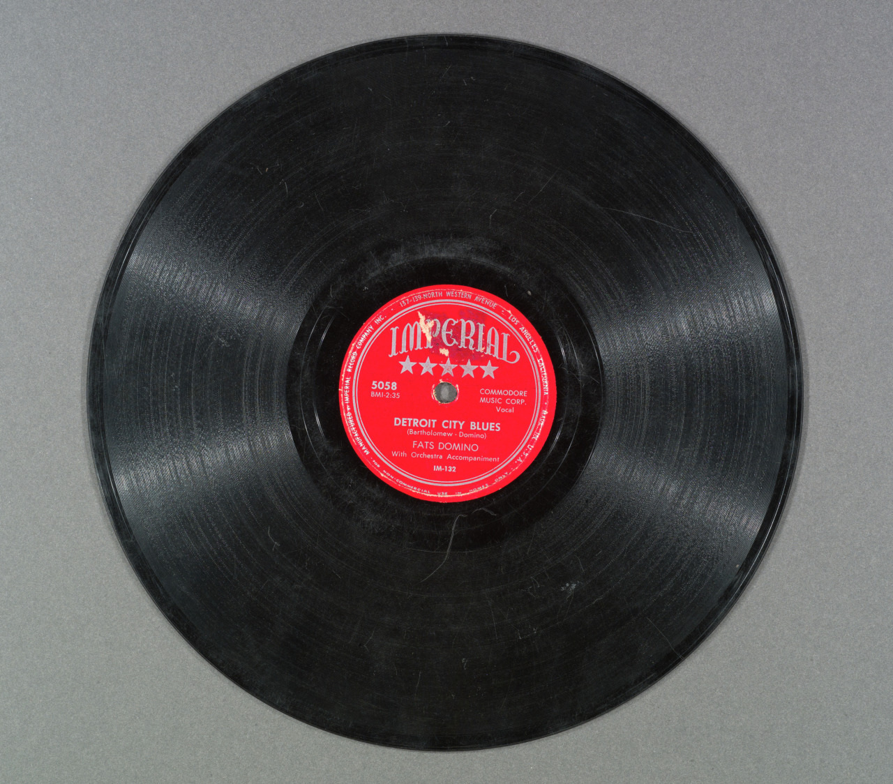 A vintage vinyl record with a red label in the center, labeled Imperial, featuring the song Detroit City Blues by Fats Domino. The record has grooves and is set against a plain gray background.