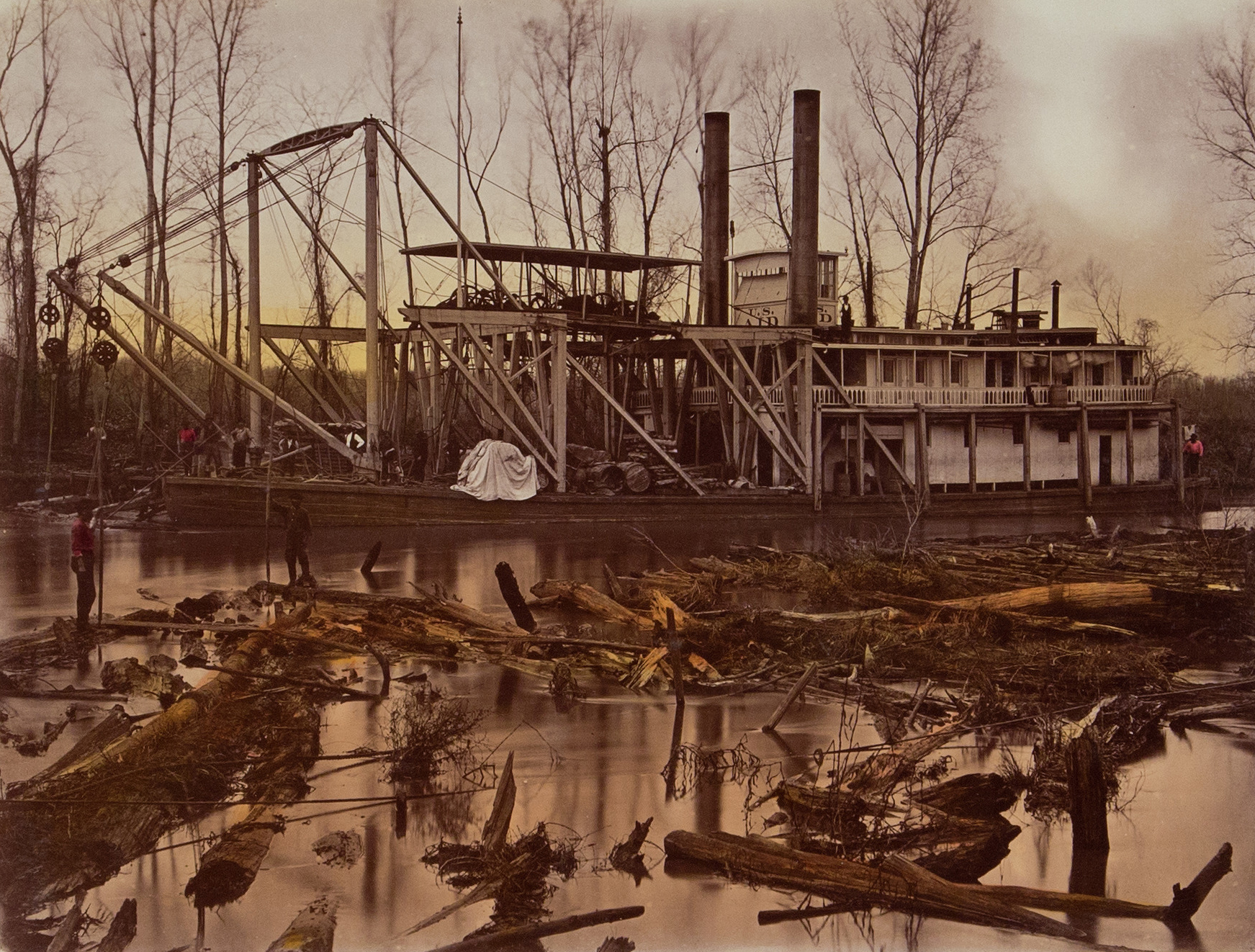 A large steamboat with cranes is docked along a riverbank surrounded by leafless trees. Logs and debris float in the water, and a few people are scattered around the boat. The sky is overcast, casting a muted light over the scene.