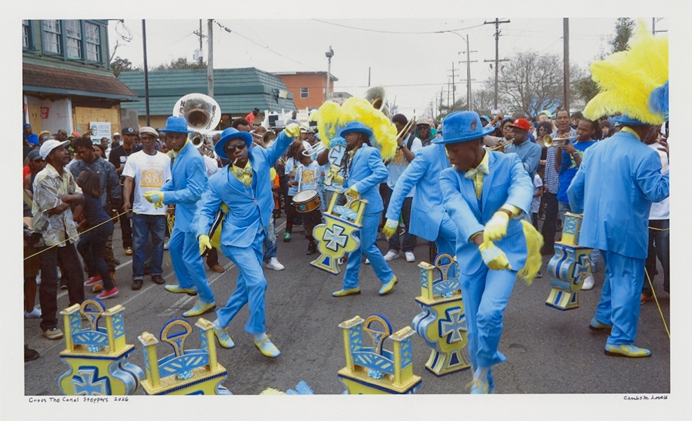 A parade featuring performers in bright blue suits, yellow feathered hats, and matching accessories. They are dancing energetically in the street as a crowd watches. Musical instruments, including tubas, are visible in the background.