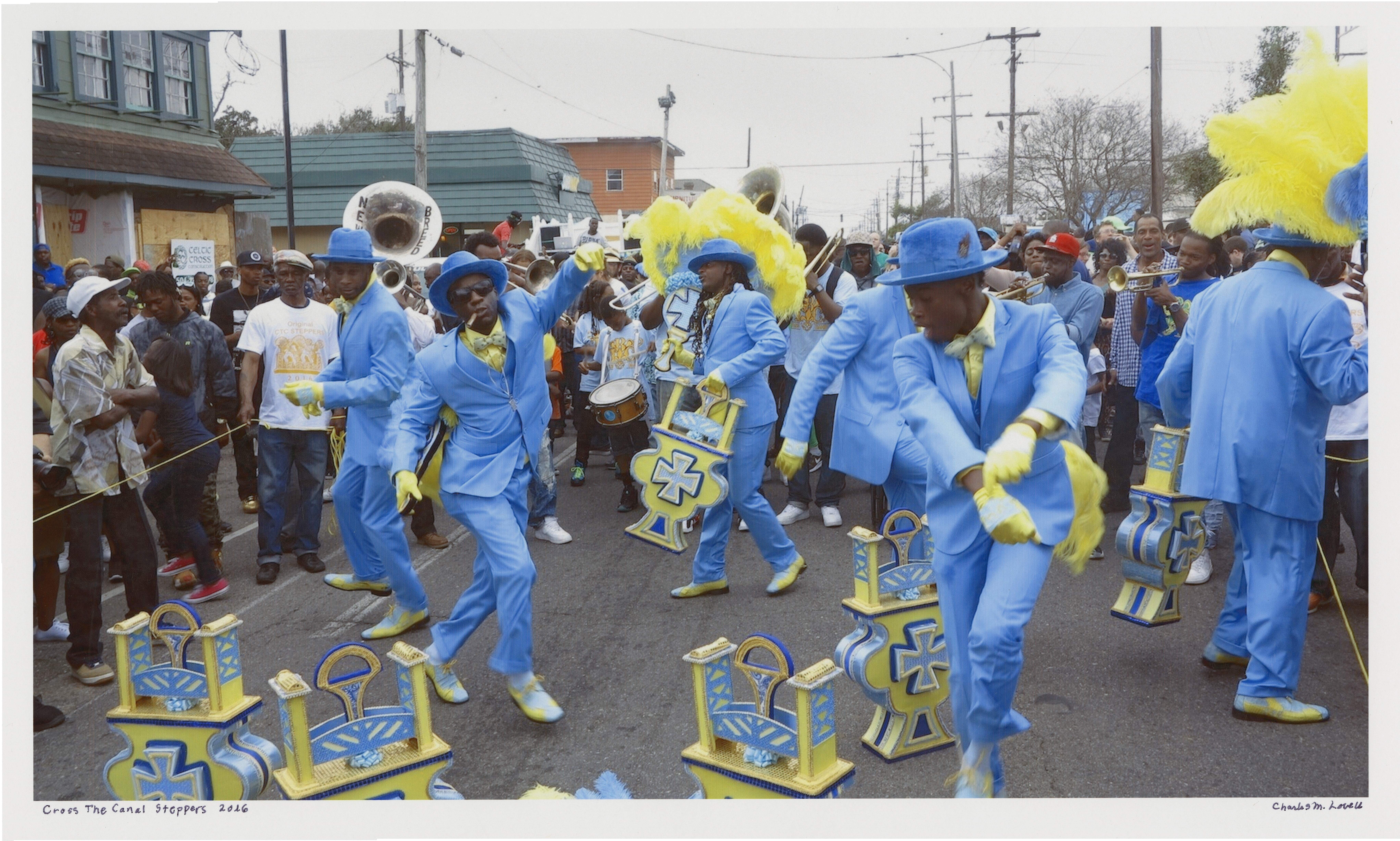 A vibrant parade scene with performers dressed in matching blue suits, hats, and yellow feather accents. They are energetically dancing and holding decorative props, surrounded by a lively crowd of onlookers on a city street.