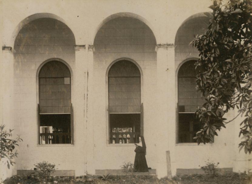 A nun stands outside a building with three large arched windows. She is facing the windows, holding a book. The building is white with some visible plants and a tree on the right. The scene has a quiet, contemplative atmosphere.