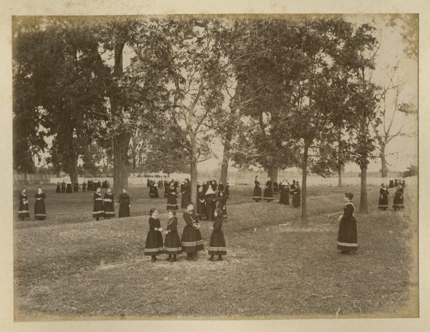 A vintage photograph of women in long, dark dresses walking and socializing in a park-like setting with large trees. Groups of two to four women are scattered across the grassy area, creating a peaceful and communal atmosphere.