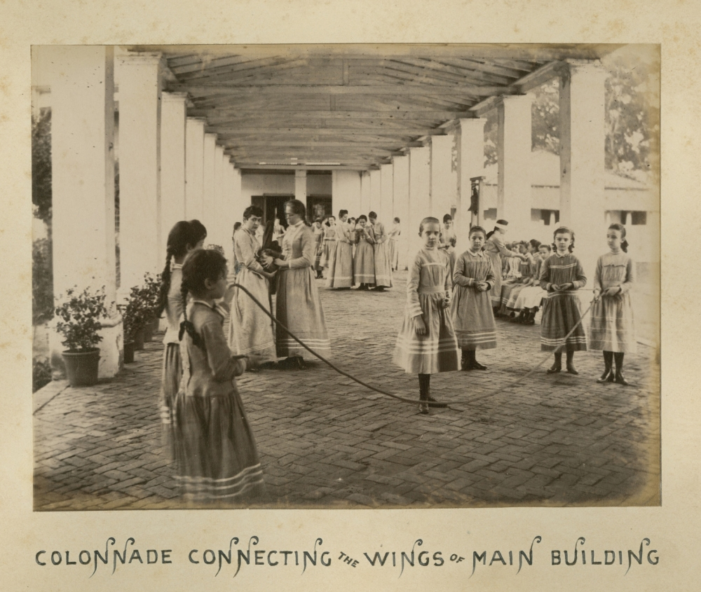 Vintage photograph of young girls in dresses skipping rope under a covered colonnade. Other girls and women observe. The setting appears to be a historical or educational institution. Signs of age are visible on the photo.