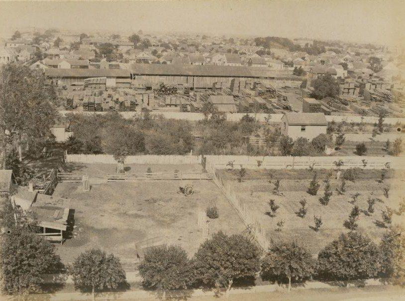 An aerial view of a historic, early 20th-century industrial area with warehouses, small trees, and open land. Residential houses are seen in the background, surrounded by a mix of greenery and fences.