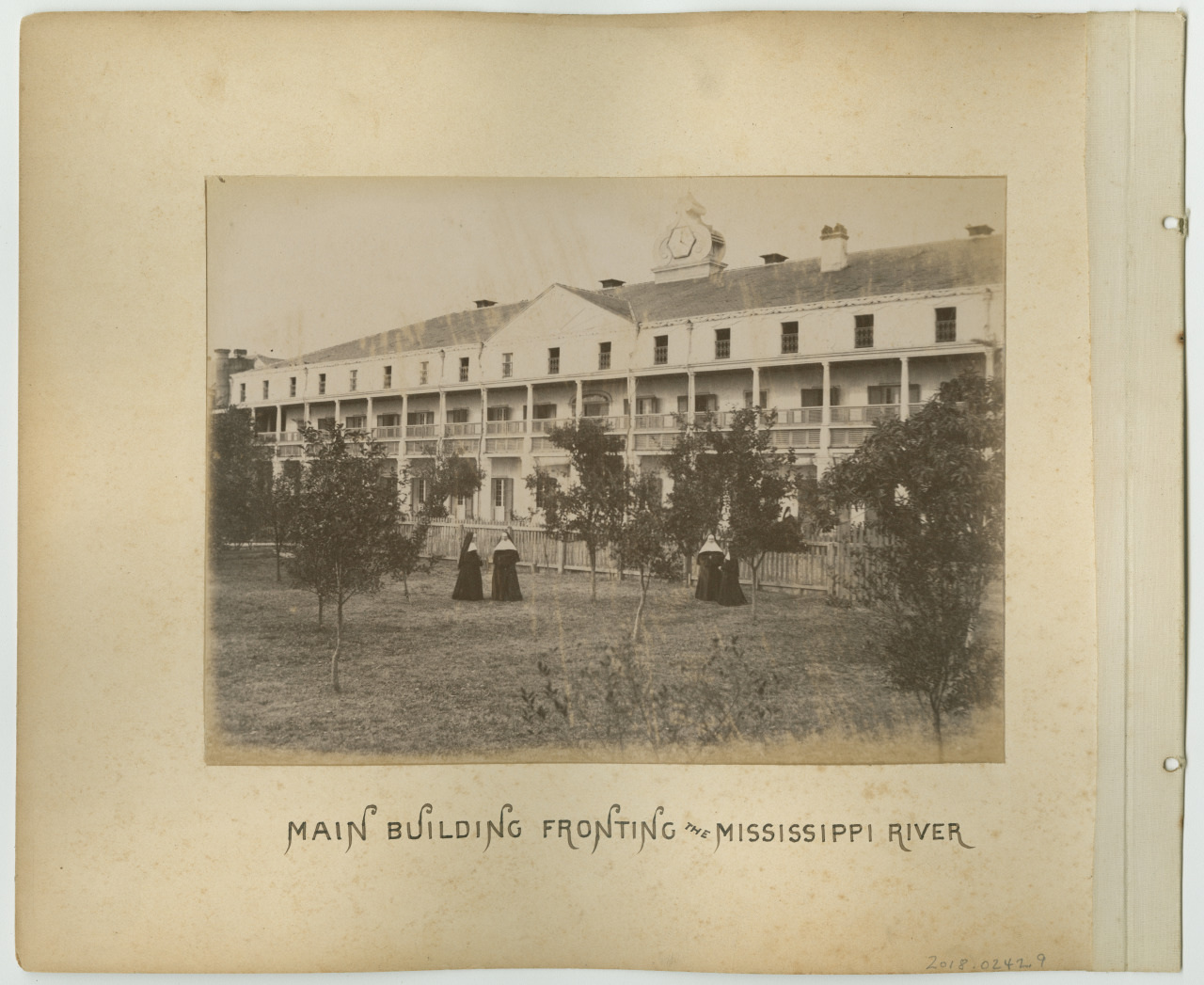 Sepia-toned image of a large multi-story building with a clock on the rooftop, facing the Mississippi River. Two people in long dresses are standing on the grass in front, surrounded by trees. Handwritten text reads Main building fronting the Mississippi River.