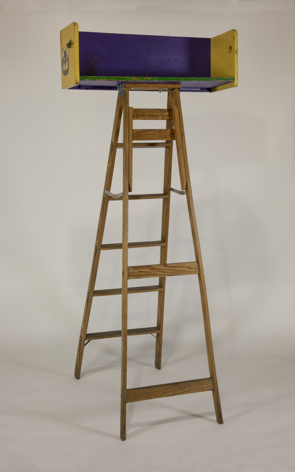 A wooden stepladder with a colorful, decorated platform on top. The sides of the platform are painted yellow and purple and feature small designs. The ladder stands on a plain, neutral background.