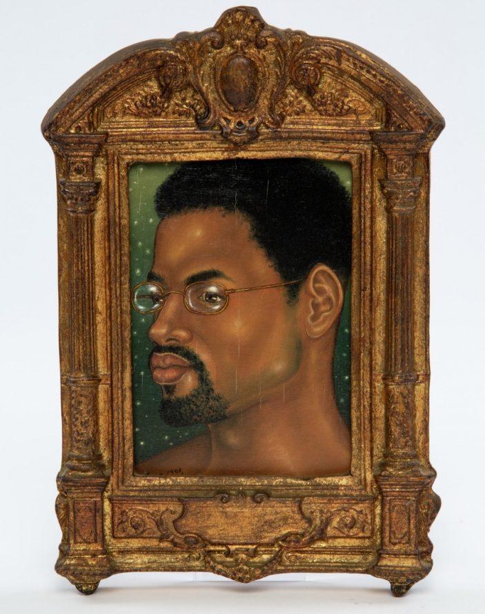 A portrait of a man with short hair and a beard, wearing glasses, set against a dark background. The artwork is housed in an ornate, antique golden frame with intricate carvings.