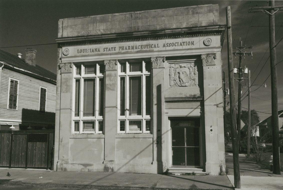 A historic stone building with large windows, featuring the inscription Louisiana State Pharmaceutical Association above the entrance. The facade includes decorative architectural details and a carved relief. Nearby are two adjacent houses and utility poles.