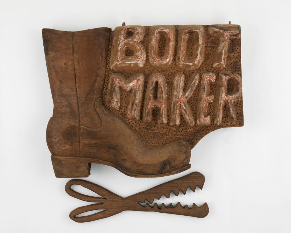A vintage wooden sign featuring a large boot with the words BOOT MAKER in raised letters. Below the boot, there are wooden cutouts resembling oversized scissors with jagged edges, all set against a white background.