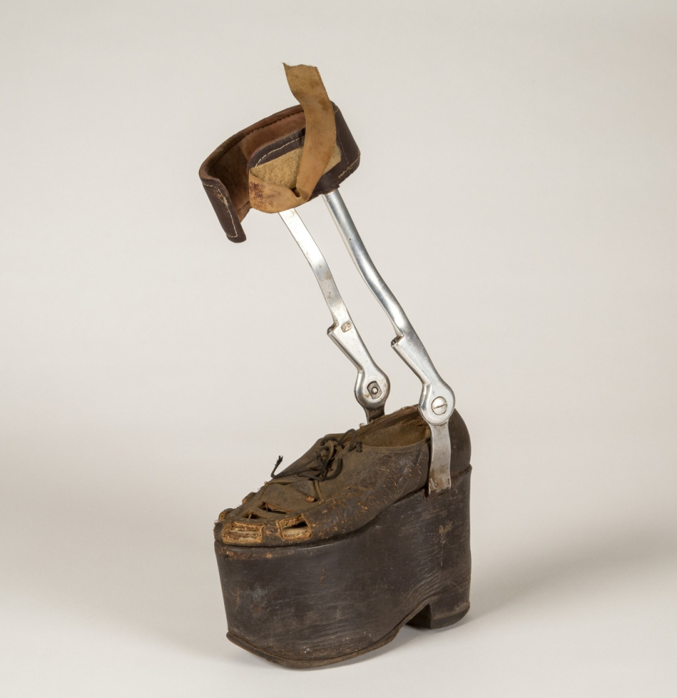 A vintage orthopedic boot with a thick, elevated sole and metal brace support. The ankle area is secured with a leather strap, and the overall design appears worn and aged, emphasizing its historical use.