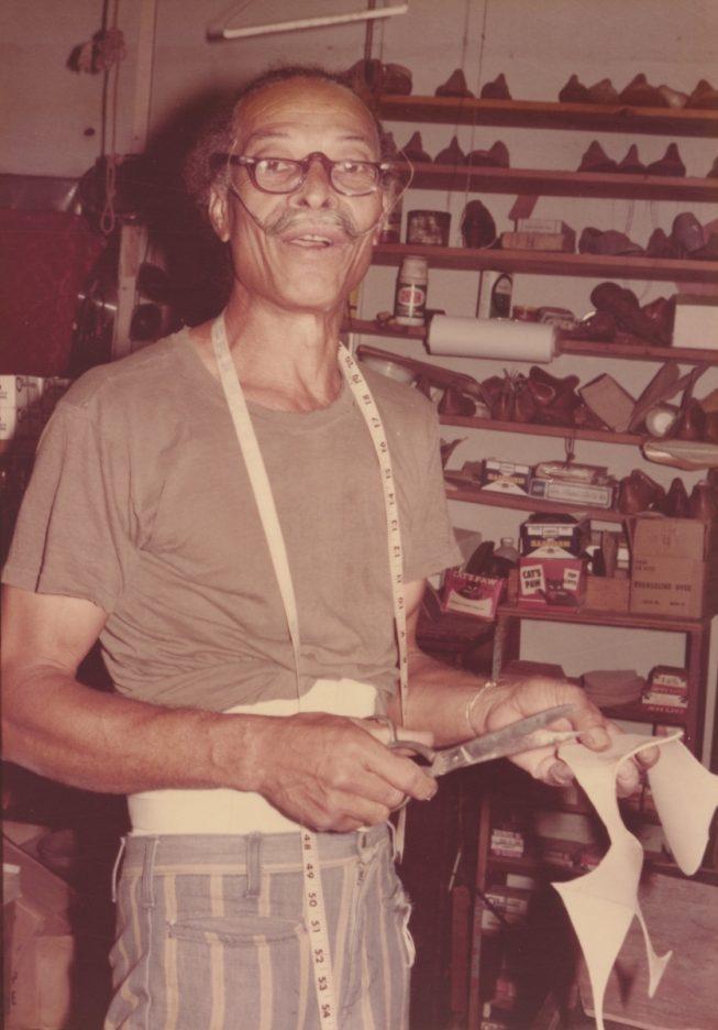 An older man with glasses stands in a workshop, holding a knife and leather material. Hes wearing a measuring tape around his neck, and shelves with shoes and materials are visible in the background.