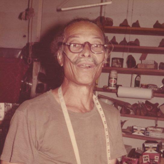 An older man with a mustache and glasses stands in a cluttered workshop. He wears a tape measure around his neck and a t-shirt. Shelves behind him hold various tools and supplies.