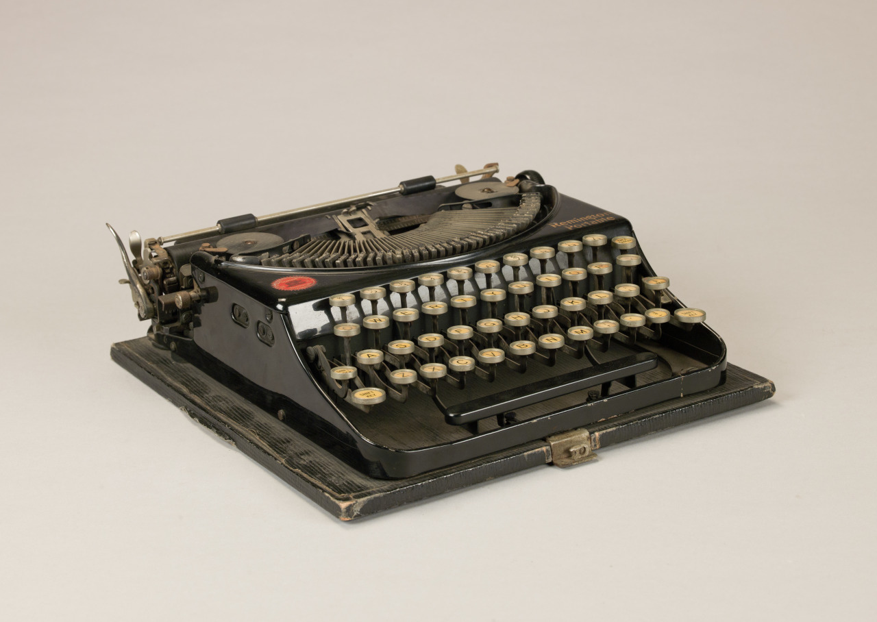 A vintage black typewriter sits on a flat surface. It features round keys and a compact design, with visible signs of wear on the edges. The background is plain, highlighting the typewriter as the main focus of the image.