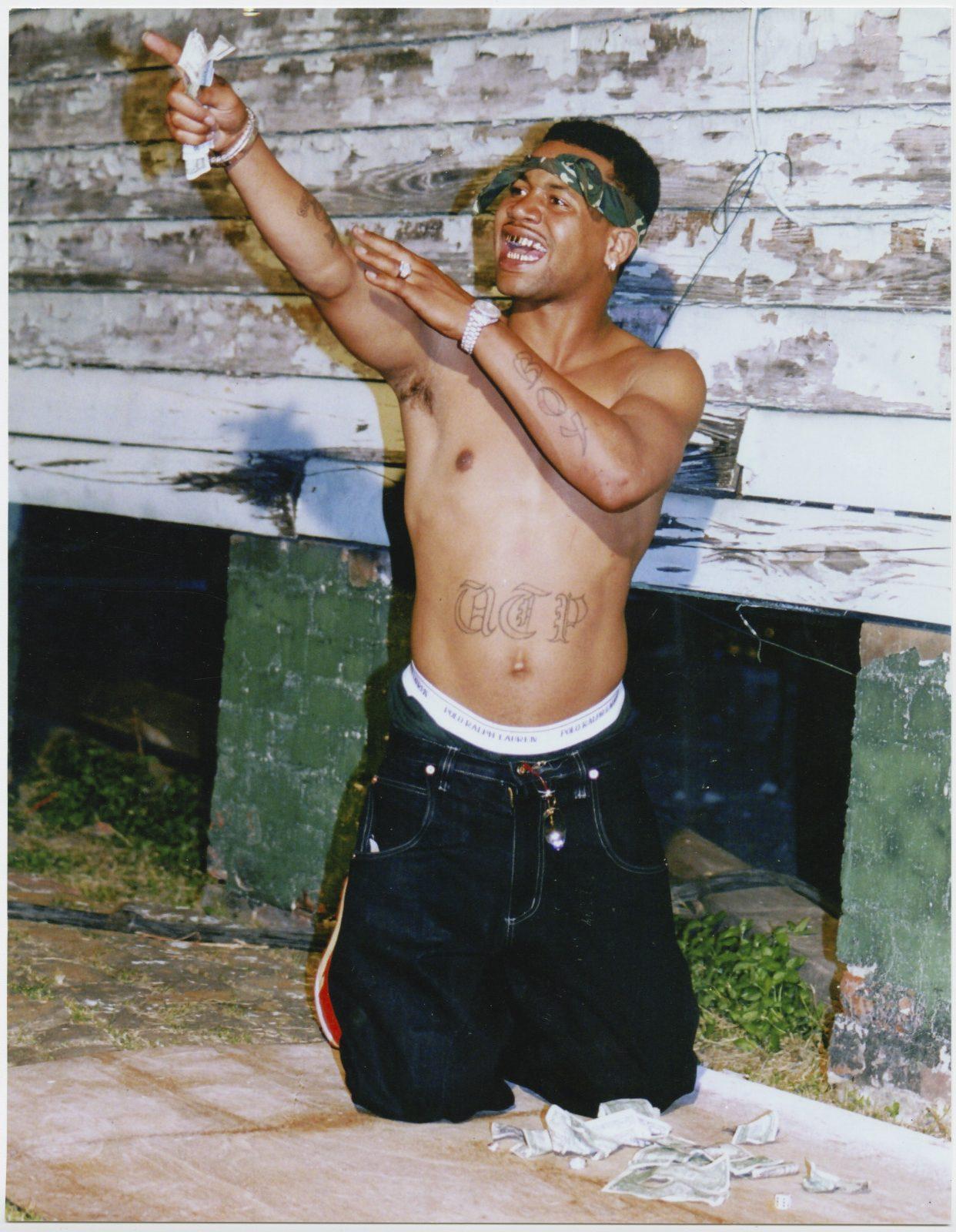 A shirtless person kneels on the ground and points upward. They have a tattoo on their chest and are surrounded by scattered money. The background features a weathered wall.