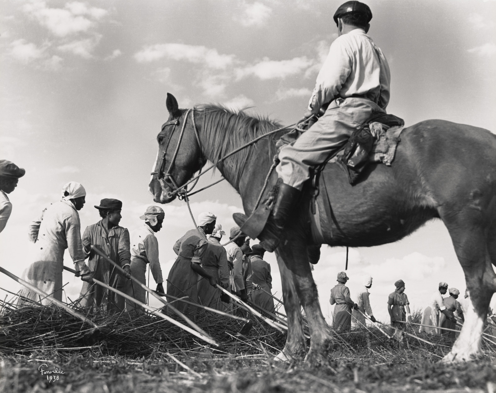 A black and white photo from 1938 shows a group of workers in a field. A person on horseback observes them. The scene depicts farming activities under a cloudy sky, with individuals dressed in work attire.