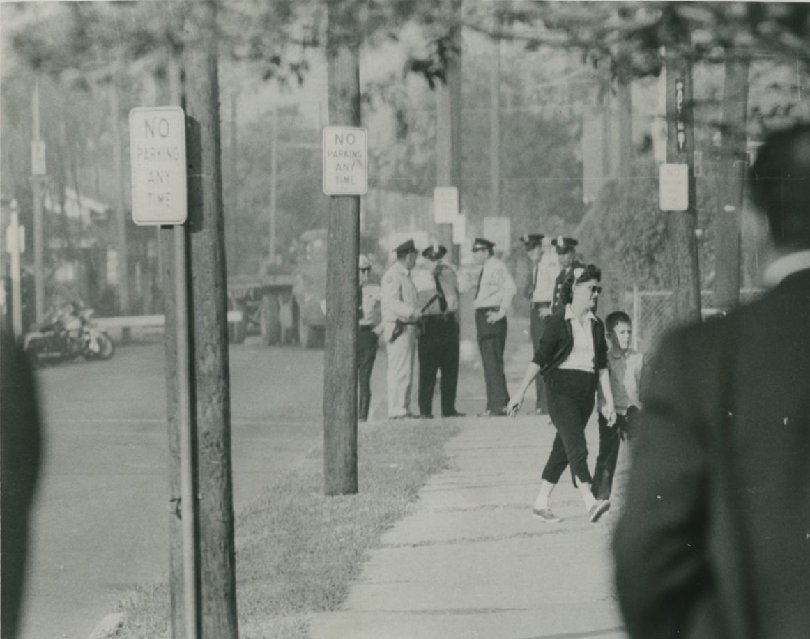 A press photograph depicting a woman walking her son to school during the third week of school integration in New Orleans, Louisiana. A group of uniformed police officers stand together on the sidewalk in the distance.