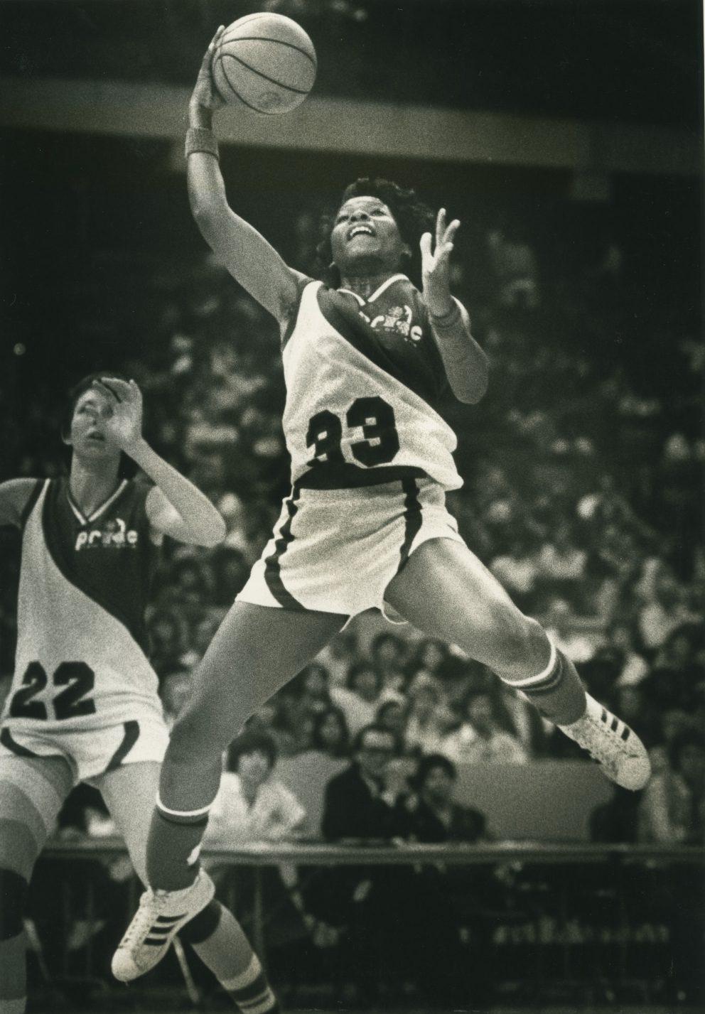 A basketball player wearing a jersey with the number 23 leaps into the air to shoot a basketball, while another player with the number 22 is nearby. The background shows a crowd watching the game.