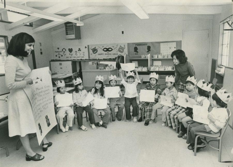 A classroom scene from the past shows two women and a group of children wearing paper crowns. The children are seated in a semicircle, holding worksheets. The room is decorated with festive wall art, including masks and the phrase Happy Mardi Gras.