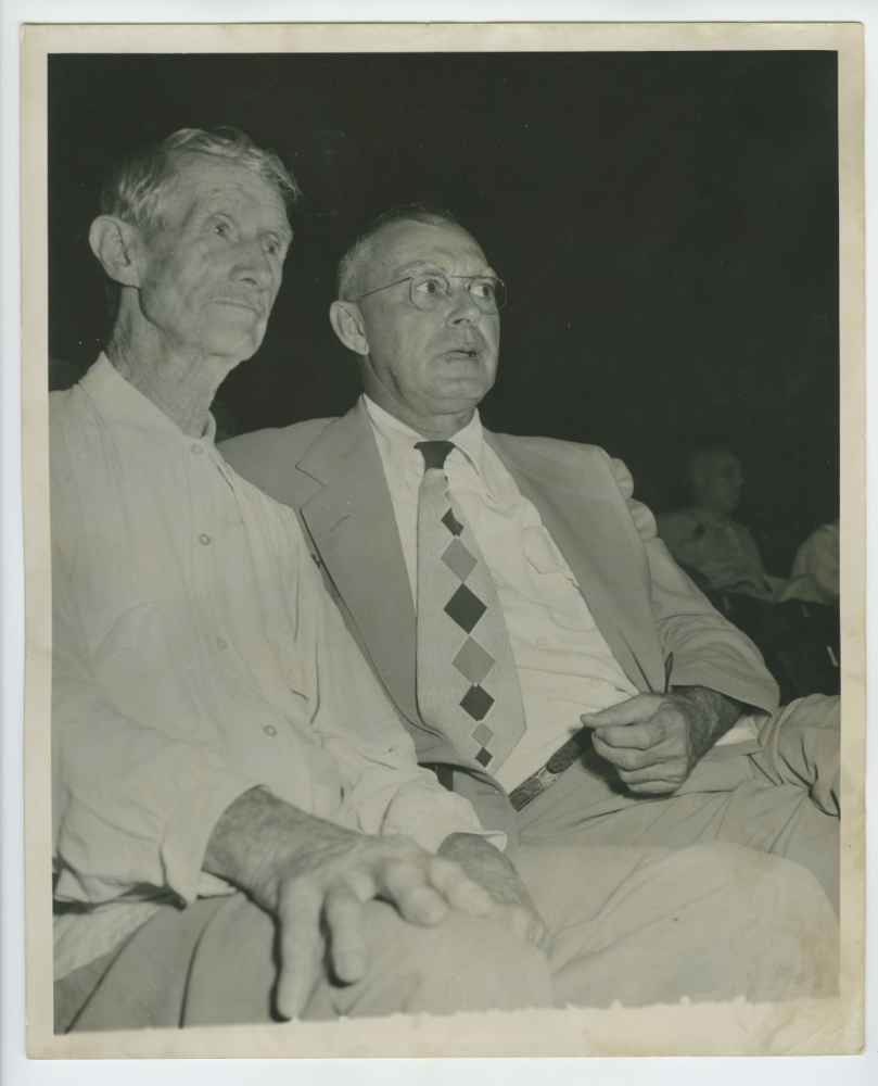 Two men sitting side by side, both with a serious expression. The man on the left is wearing a light-colored shirt, and the man on the right is in a suit with a patterned tie, holding a cigar. The background is dimly lit. The photo appears vintage.