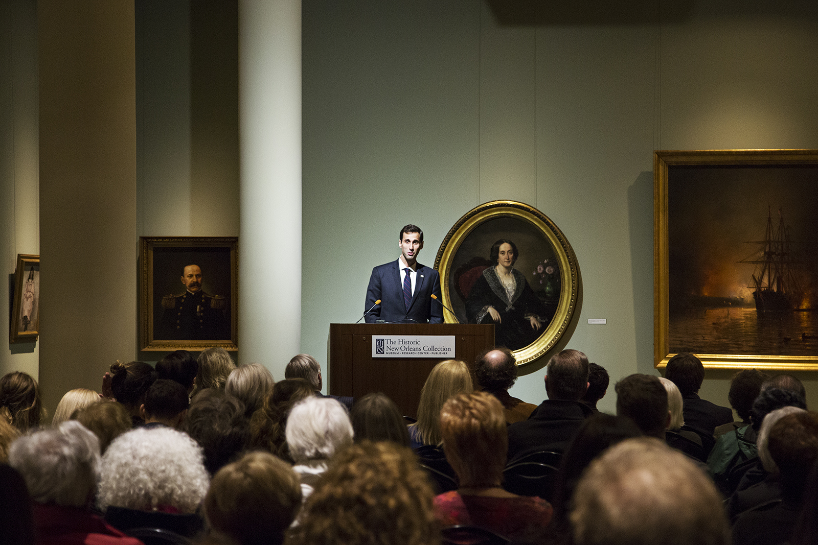 A person stands speaking at a podium in a dimly lit art gallery filled with people. The walls are adorned with portraits, and the audience is seated facing the speaker.