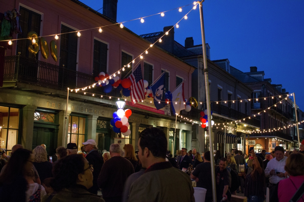 A bustling street festival scene at dusk, with string lights overhead and lively crowd mingling. American and other flags hang above, and colorful balloons add festive flair. Historic buildings line the street under a deepening blue sky.