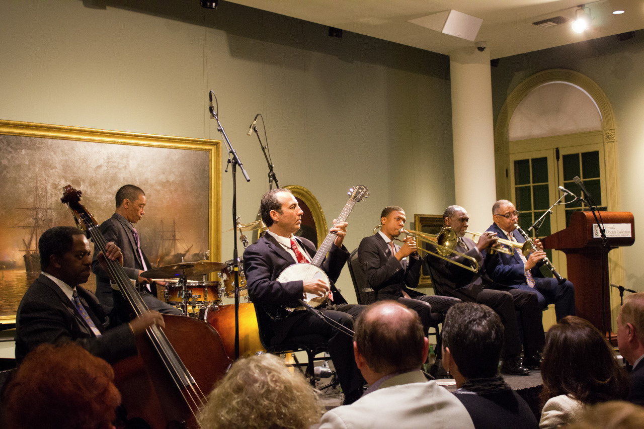 A jazz band performs in a gallery, featuring musicians on banjo, double bass, drums, trumpets, and clarinet. They are seated in front of a painting, with an audience watching. The room has soft lighting and elegant decor.