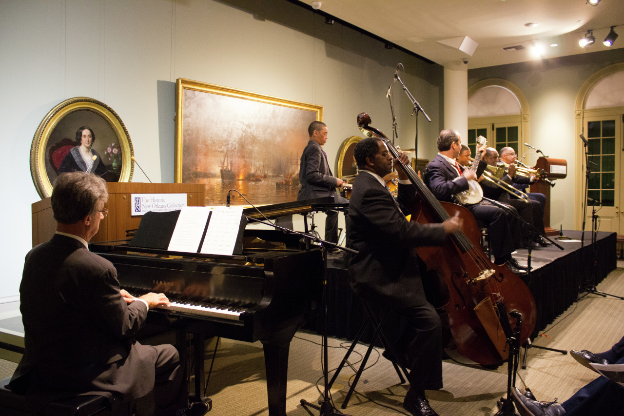 A jazz band performs on stage with musicians playing the piano, double bass, saxophone, trumpet, and trombone. They are in a dimly lit room with framed artwork on the walls and a small seated audience.