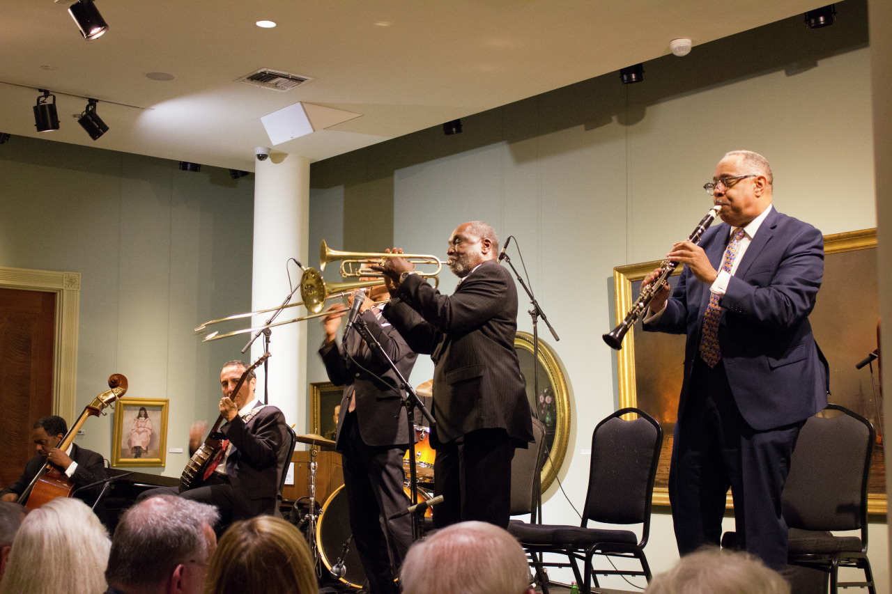 A jazz band performs on stage. Five musicians are playing instruments, including a clarinet and a trombone. They are dressed in suits, and an audience watches attentively. Portraits hang on the green walls behind them.