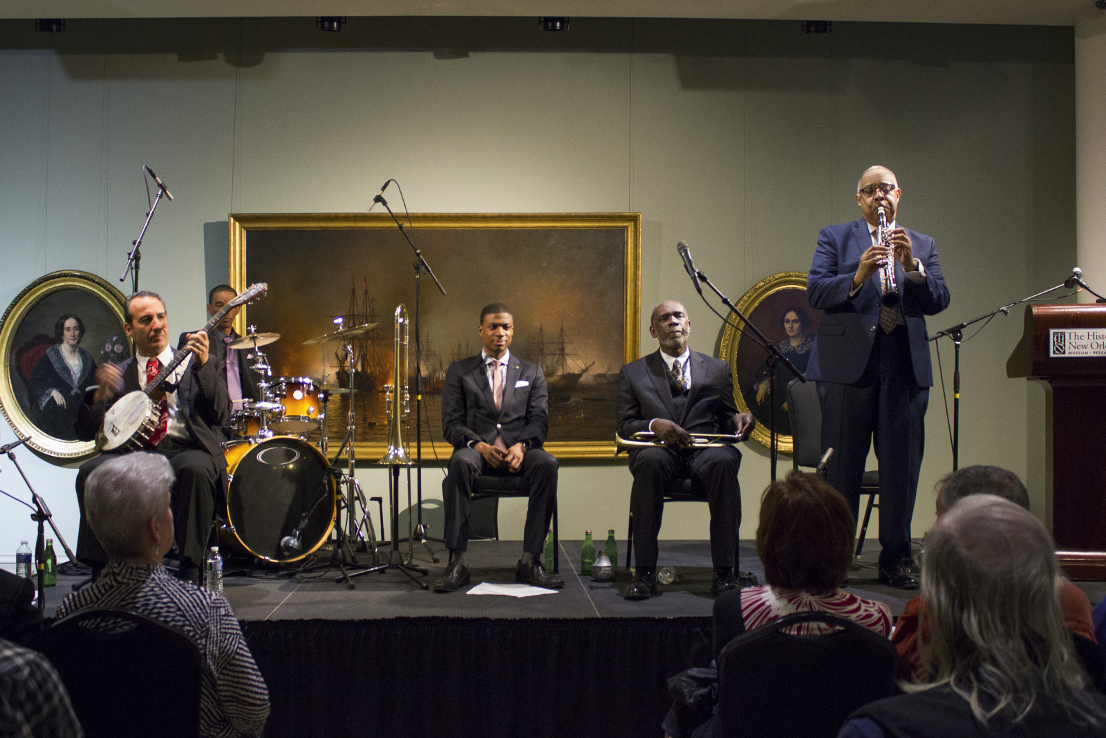 A jazz band performs on stage with four musicians. One plays the clarinet while standing, and the others sit holding instruments. Behind them are large framed paintings. An audience watches in front of the stage.