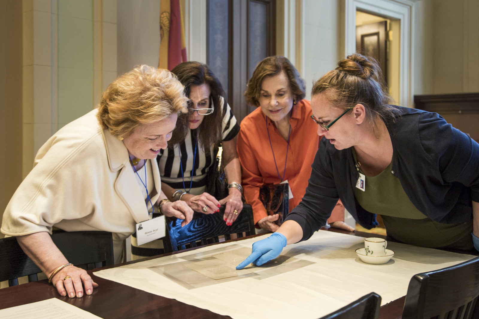 Four women gather around a table examining a historical document. One woman wearing gloves points at the document. They appear engaged in discussion. A small teacup is visible on the table. The setting is a well-lit office or conference room.