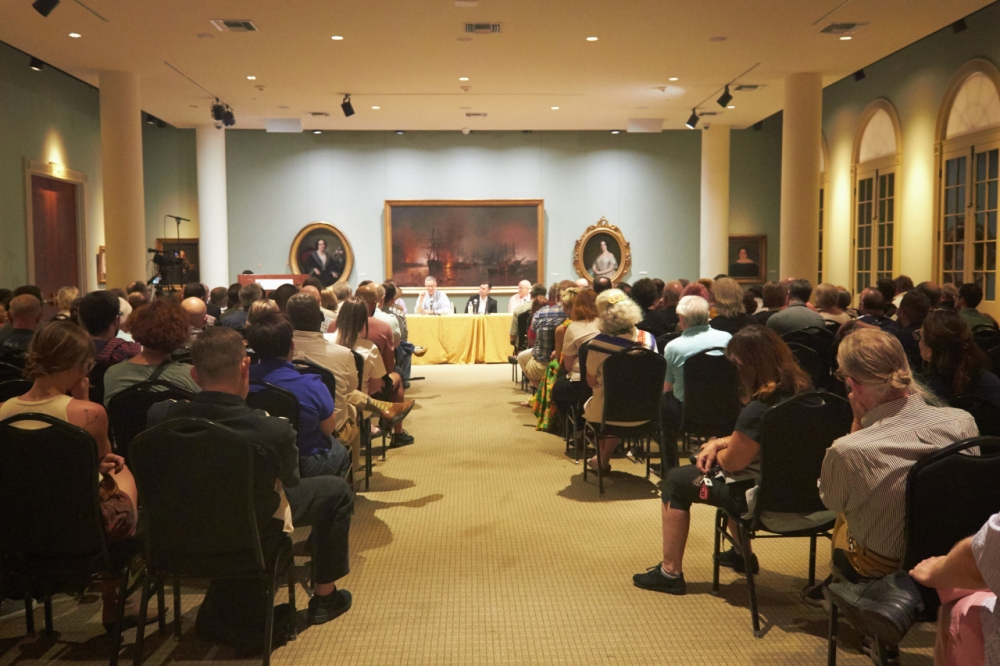 A large audience sits in rows of chairs facing a panel of speakers in a formal room. The room has green walls, art, and columns. The panel members sit at a table covered with a yellow cloth, engaged in discussion.