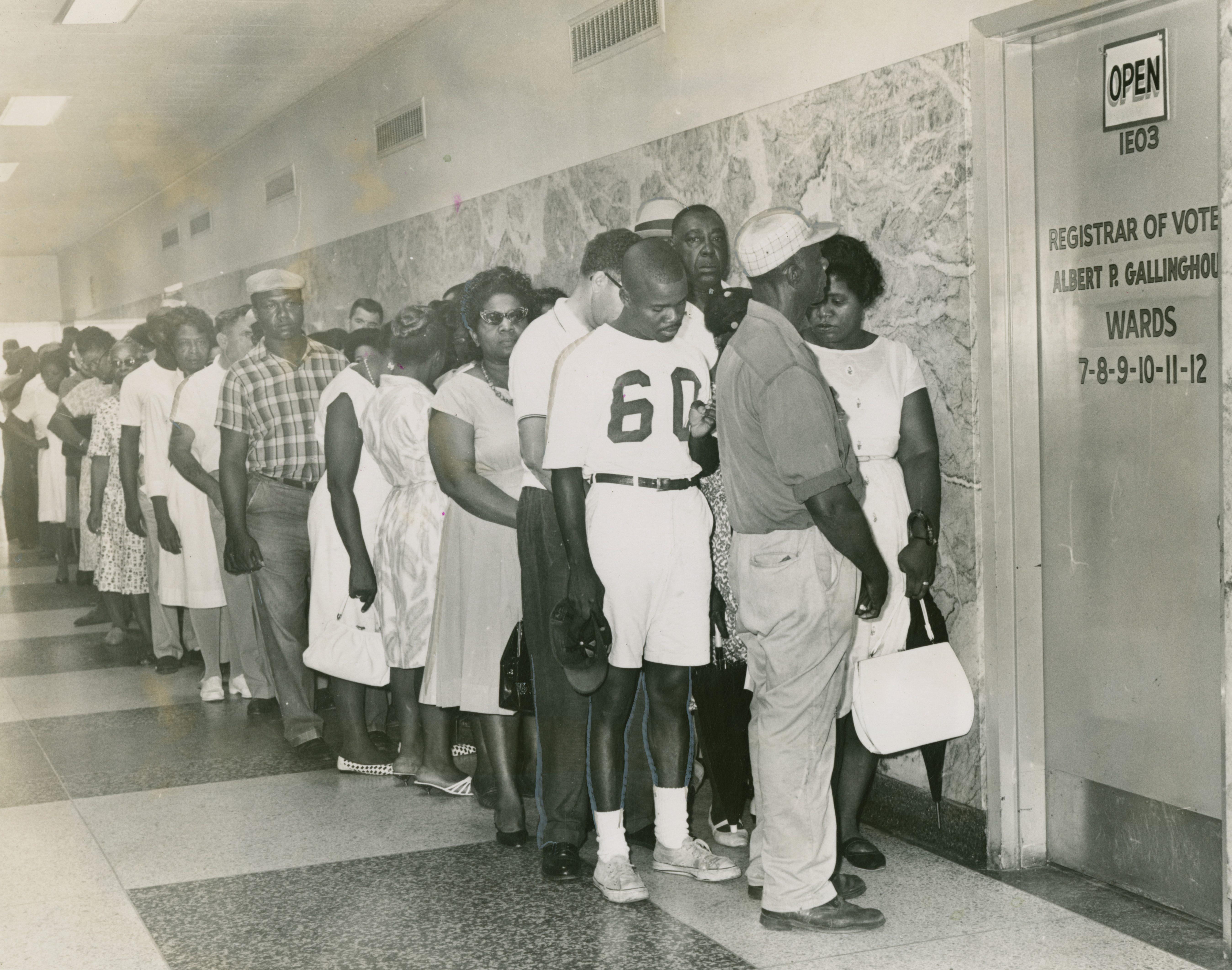 Black and white photo of a diverse group of people standing in a long line in a hallway. They are waiting outside a door labeled Registrar of Voters with ward numbers listed. The atmosphere is orderly and focused.
