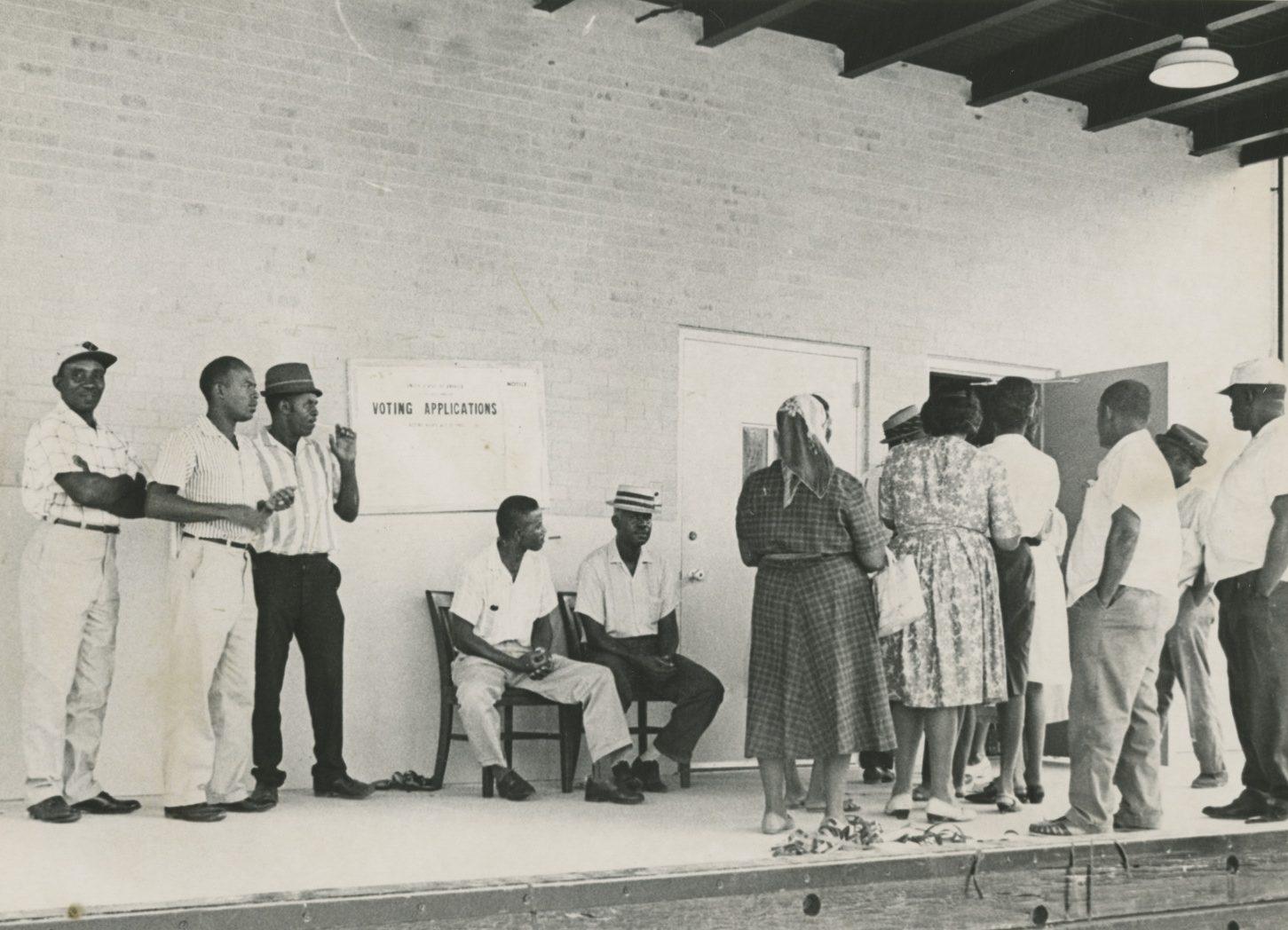 Black and white photo of people in line for voting registration outside a building. Two men are seated near a Voting Applications sign, and several individuals are standing in line, some wearing hats and summer clothing.