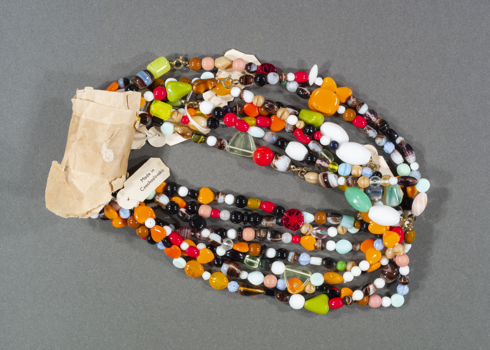 A tangled pile of colorful beaded necklaces with various shapes and sizes of beads, including red, orange, green, and white. There is a small beige tag and a crumpled piece of brown paper nearby, all on a gray background.