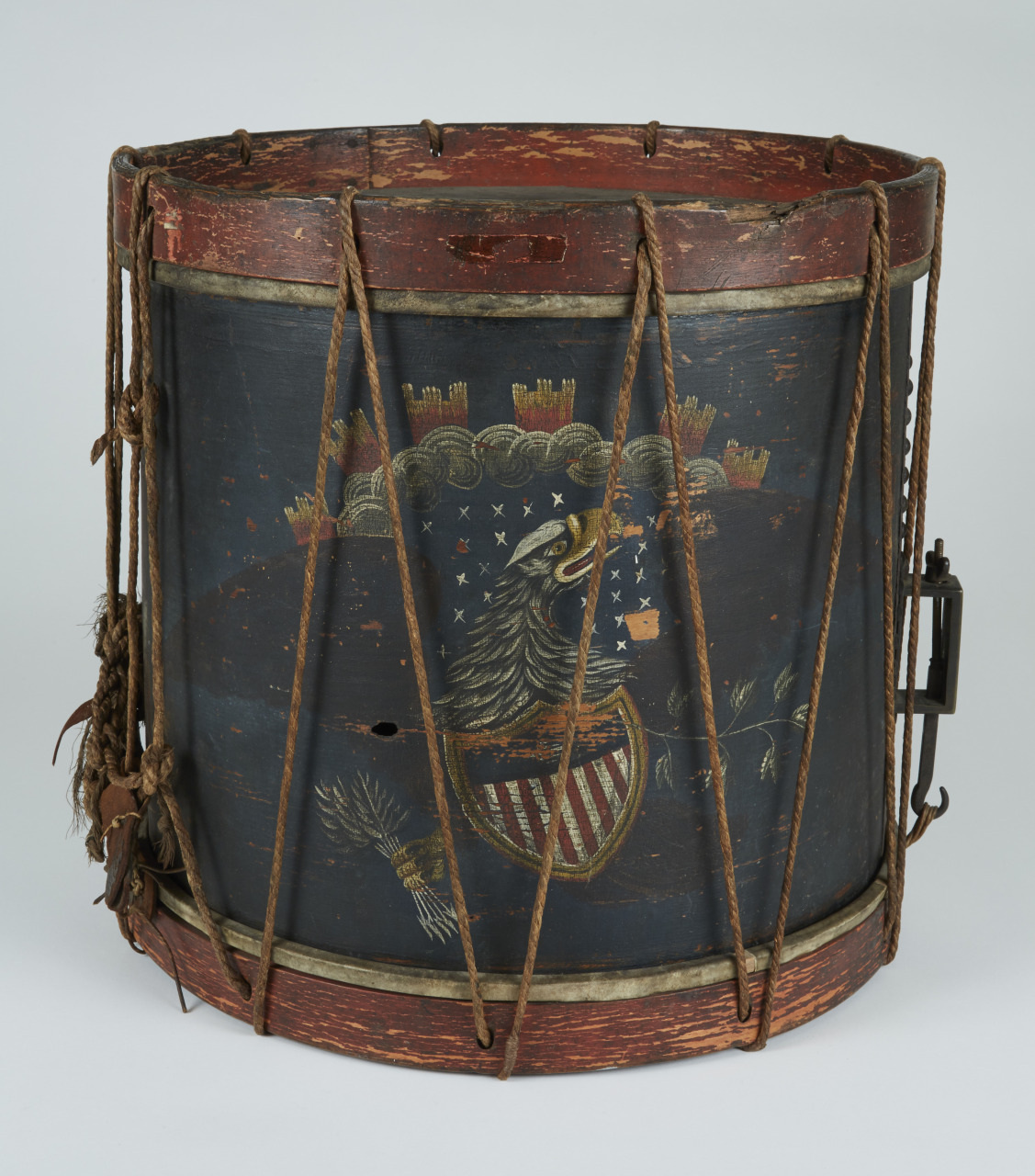 A vintage military drum featuring an eagle with a U.S. shield, surrounded by stars and decorative elements. The drum has worn red and blue paint, with rope tensioning and wooden rims, showing signs of age and use.