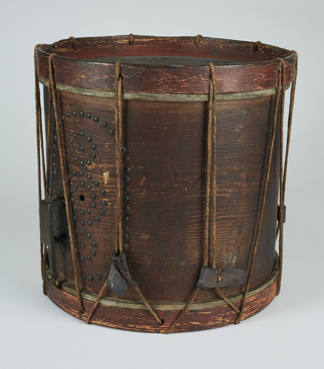 A vintage wooden drum with a dark cylindrical body and rope-tensioned skins. The drum is adorned with metal accents and displays signs of wear, giving it a rustic, aged appearance.