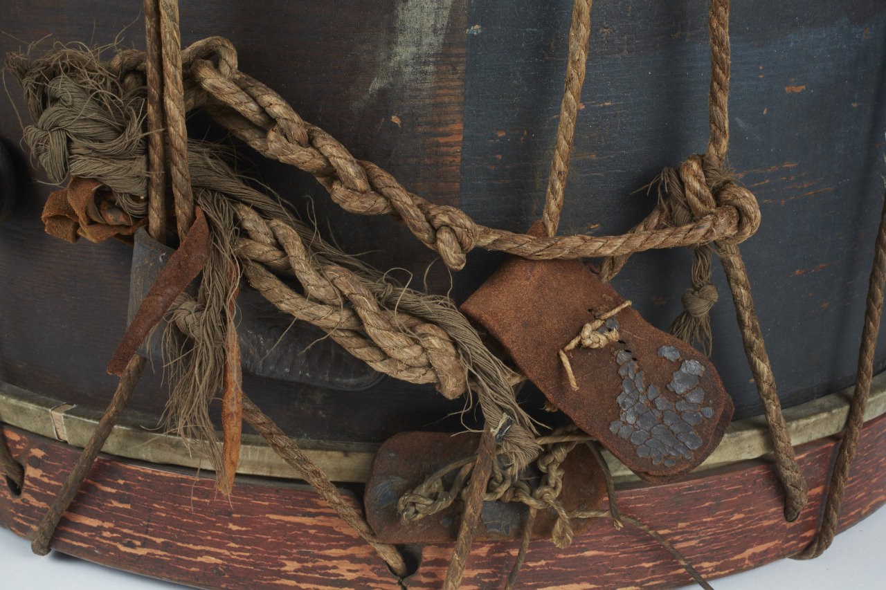 Close-up of a weathered, dark brown drum with frayed ropes and leather straps wrapped around it. The wood shows signs of age with a chipped red-painted base, giving a rustic, vintage appearance.
