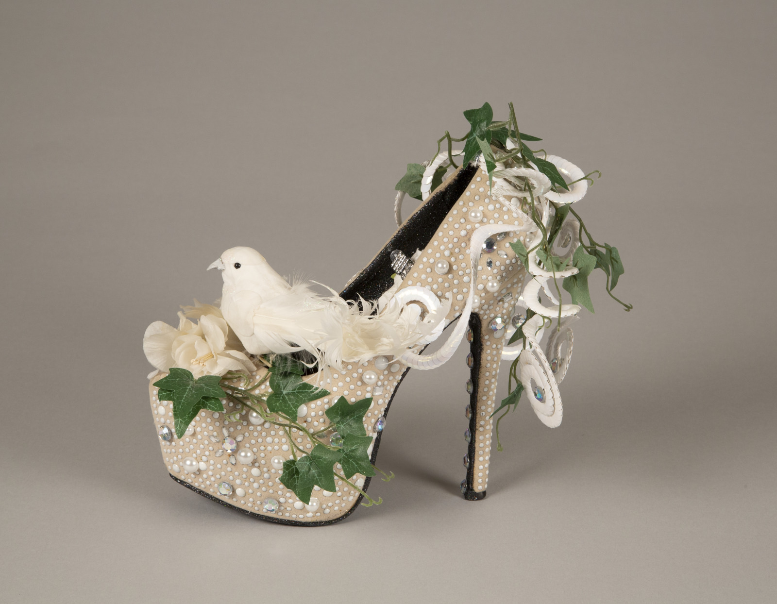 A high-heeled shoe adorned with pearls, sequins, and a faux white bird. Decorative flowers and green ivy cascade over the shoe, giving it an elaborate, nature-inspired design. The background is a neutral gray.