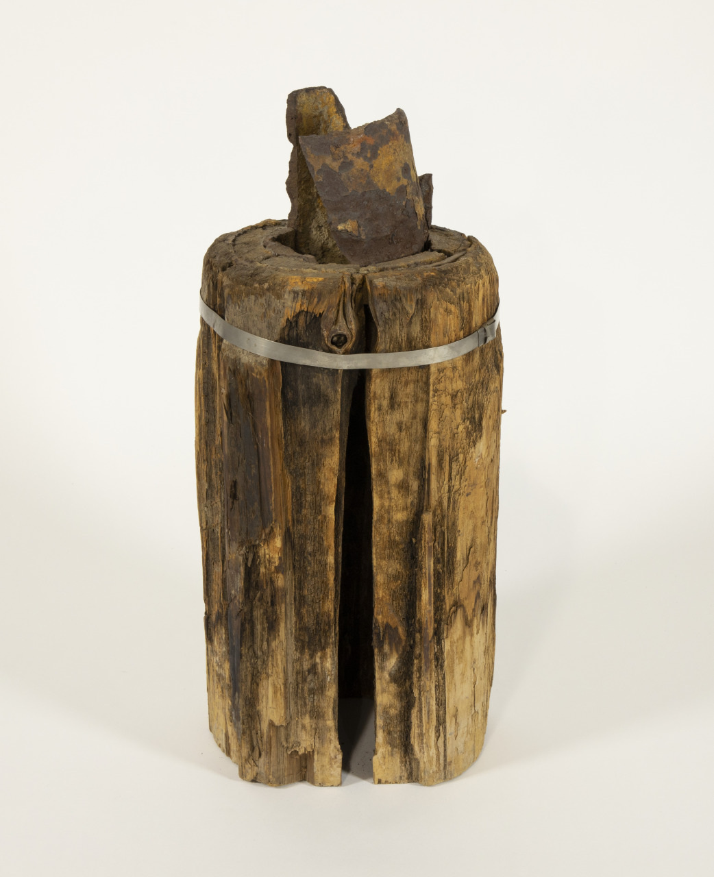 A rustic, cylindrical wooden stump with a metal band around it, featuring a rough, jagged piece of iron protruding from the top, displayed against a plain white background.