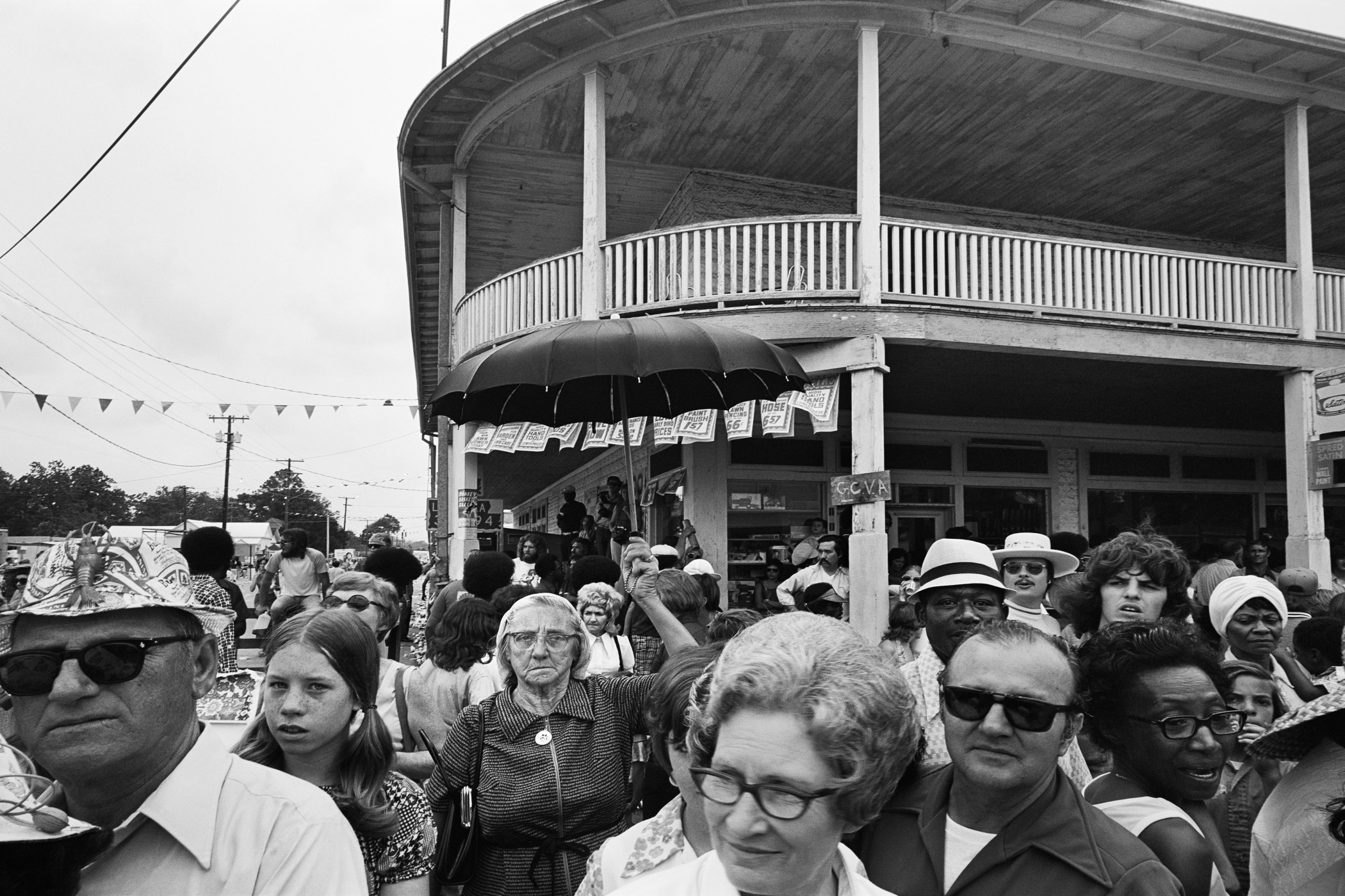 A diverse crowd of people gathered outdoors in front of a large, two-story building with a wraparound porch. Some individuals wear hats and sunglasses. An umbrella is held above one persons head. Bunting hangs from the porch.