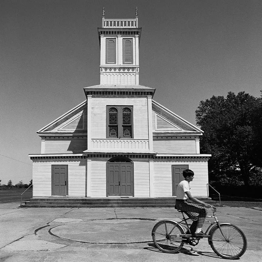 A person rides a bicycle in front of a large, white wooden church with a tall steeple and multiple doors. The sky is clear, and trees are visible to the right. The architecture is symmetrical and detailed.