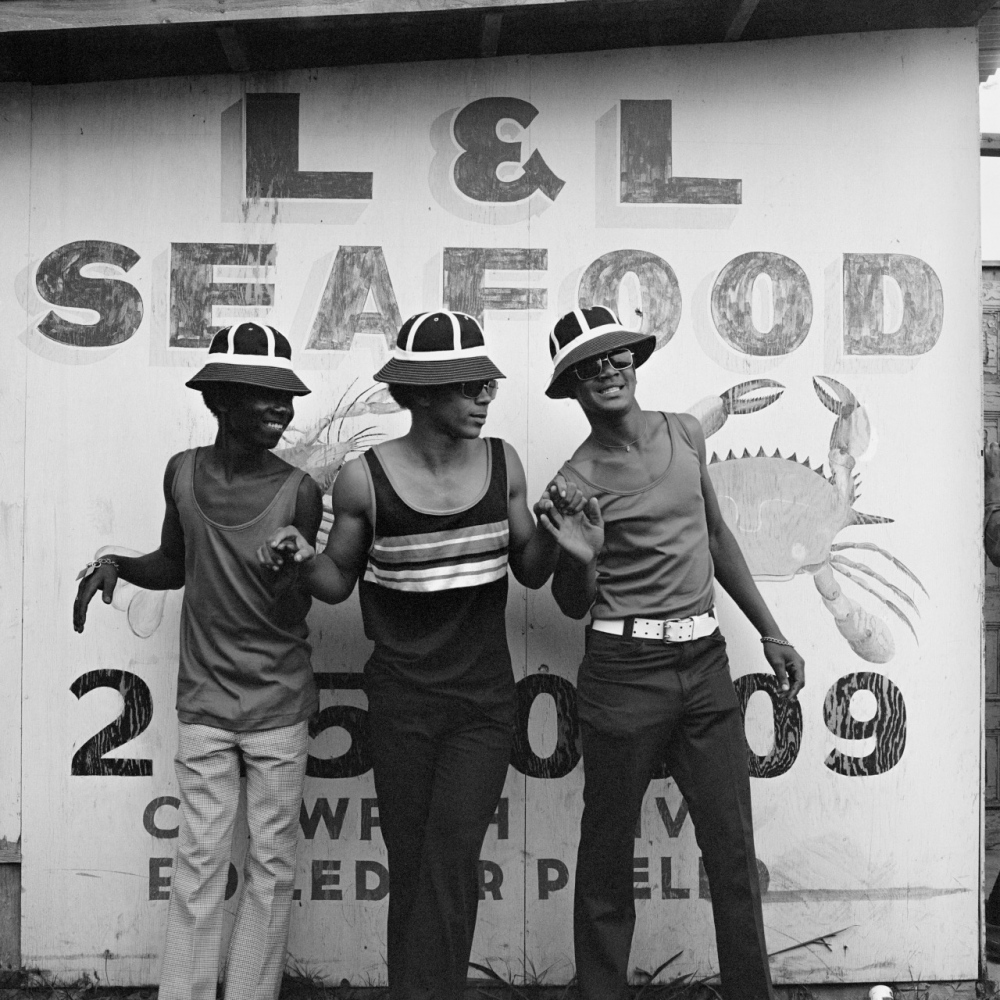 Three people wearing hats and stylish clothing pose joyfully in front of an L & L Seafood sign with a crab illustration.