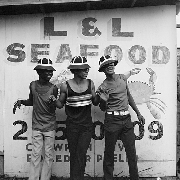Three men in stylish hats and clothing pose playfully in front of an L&L Seafood market sign with a crab design. The image is in black and white.