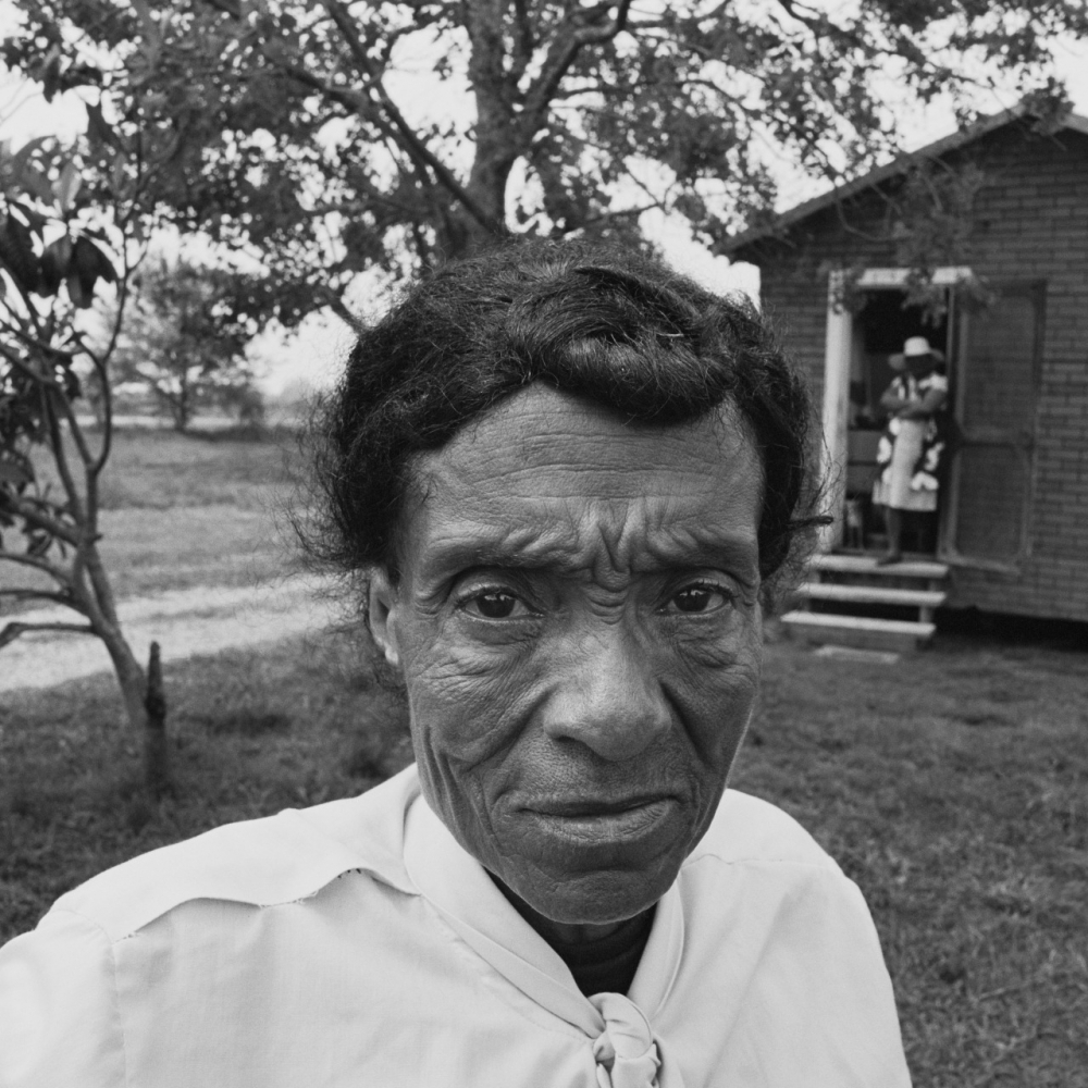 Black and white photo of a person with a serious expression standing outdoors. A small building and trees are in the background. Another person is seen at the entrance of the building. The scene appears calm and rural.