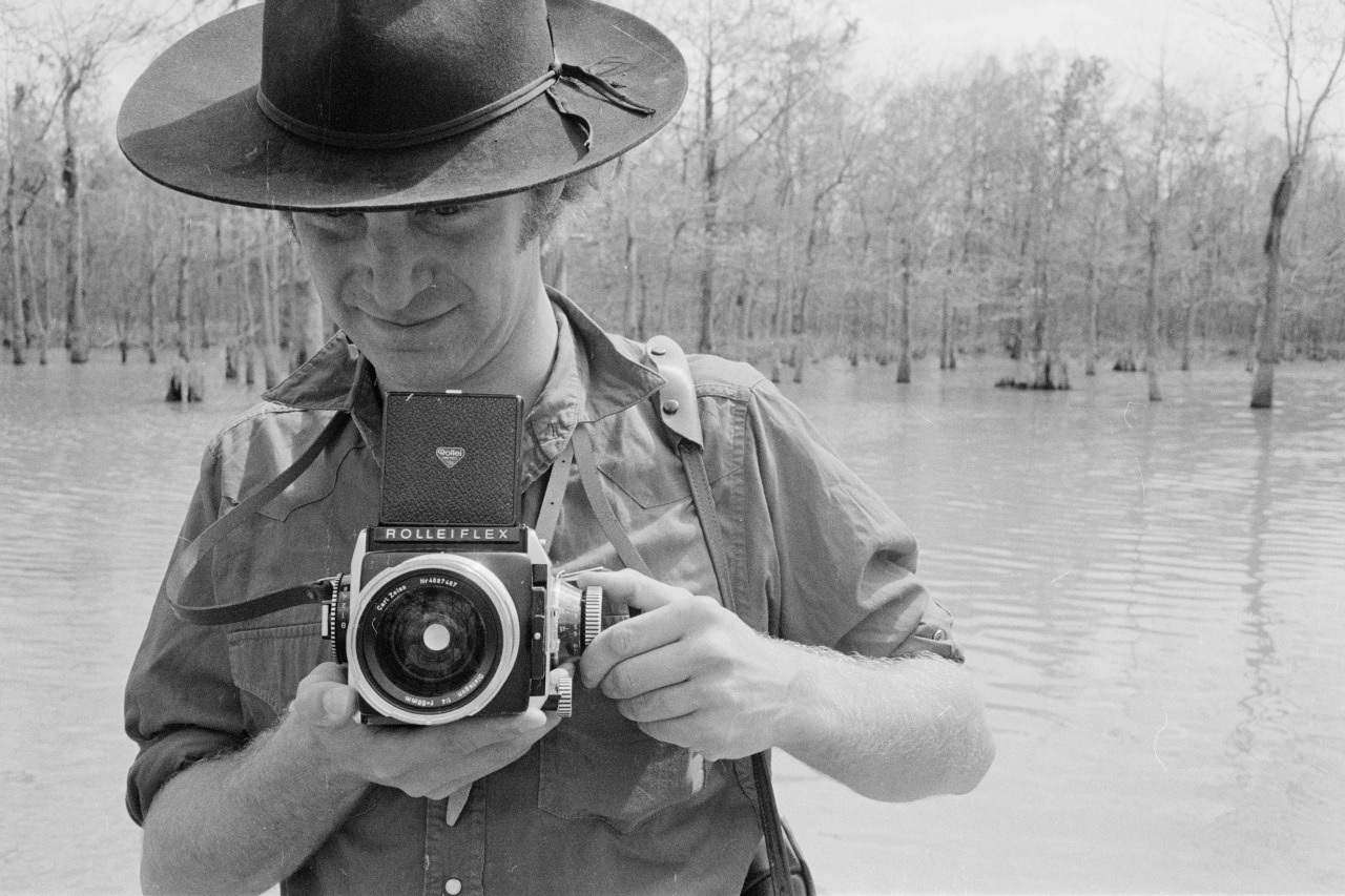 A person wearing a wide-brimmed hat holds a Rolleiflex camera, standing near a calm body of water with leafless trees in the background. The scene appears to be outdoors in a swamp or marshy area.