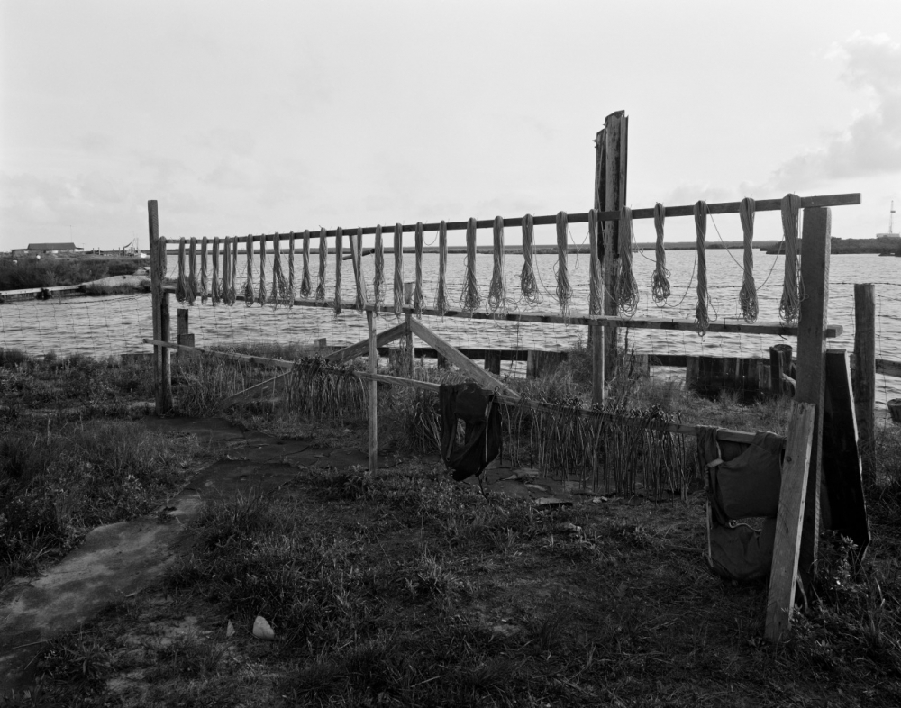 Black and white image of a wooden drying rack with ropes hanging, set near a body of water. Grass and equipment are scattered around the foreground, with an overcast sky in the background.
