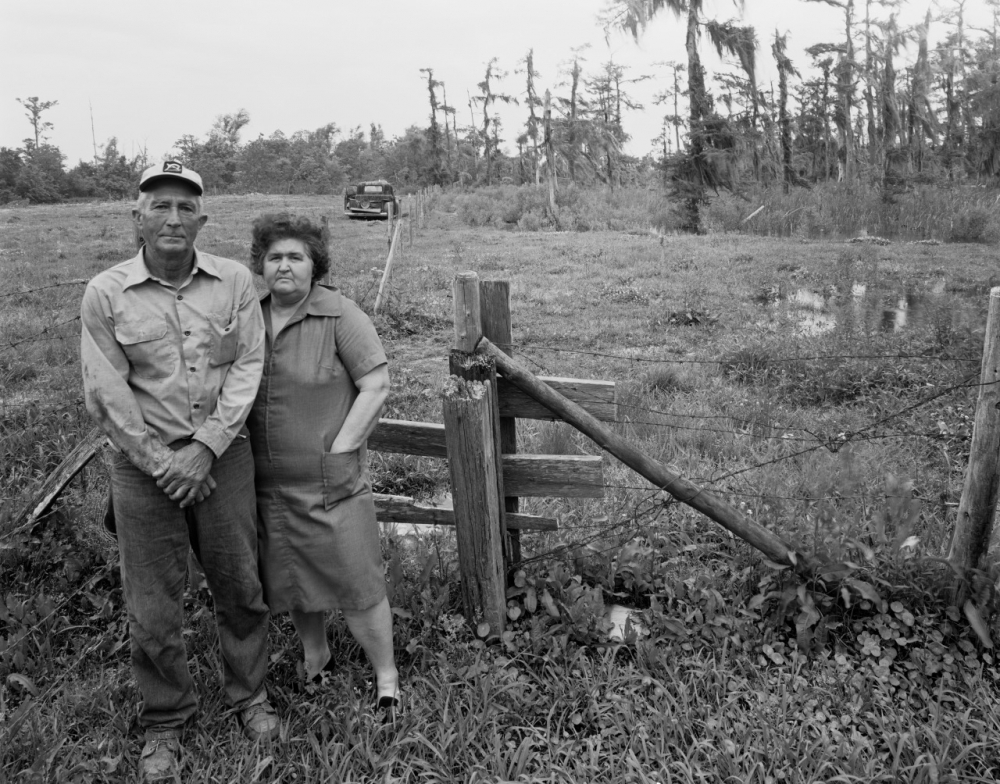 An older man in a cap and a woman in a dress stand in a grassy field by a wooden fence, with a calm pond and trees in the background. An old car is parked at a distance under a cloudy sky.