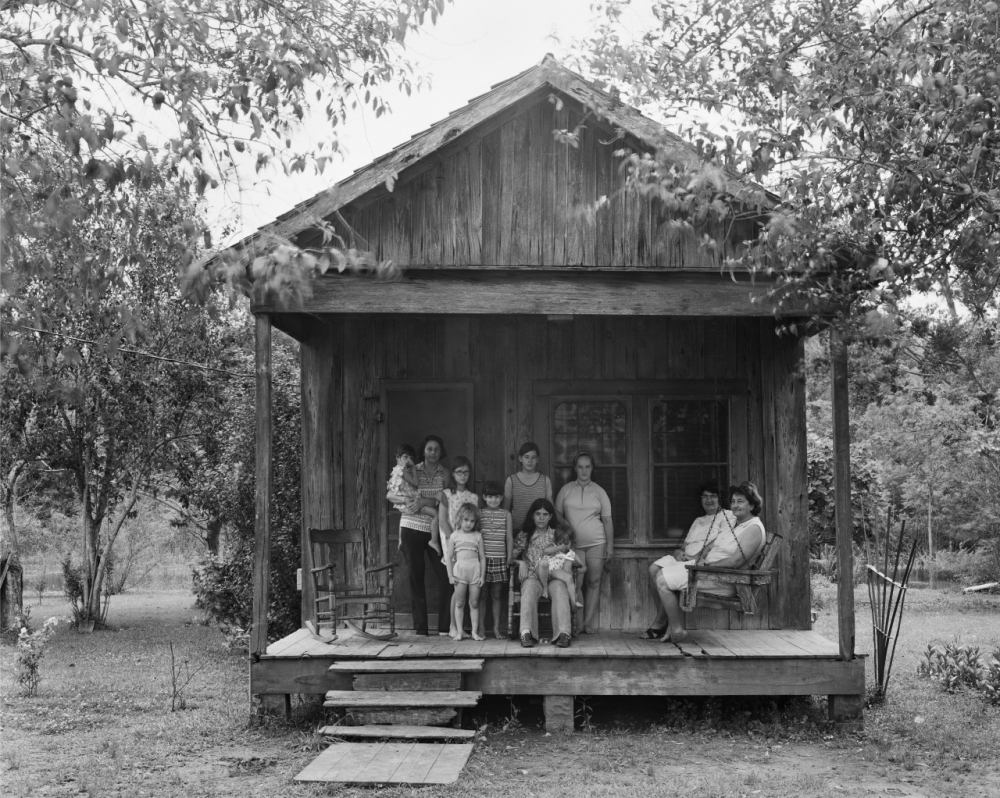 A black and white photo of ten people on the porch of a rustic wooden house, surrounded by trees. The group consists of adults and children, with some sitting and others standing, creating a relaxed and communal atmosphere.