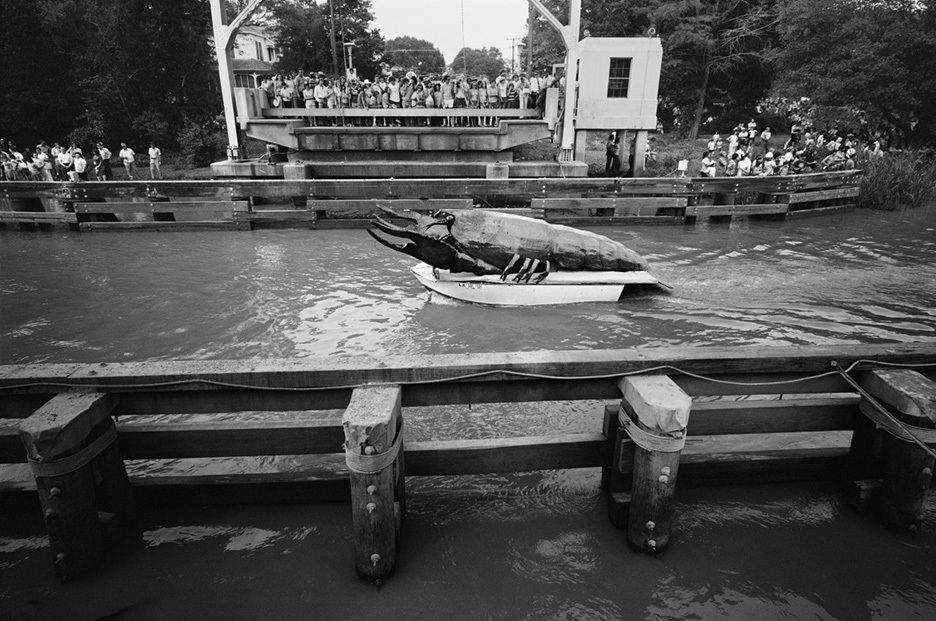 A boat decorated with a large turtle floats on a canal, surrounded by a crowd of people watching from a bridge and the banks. The scene appears festive, with spectators engaged in the event.