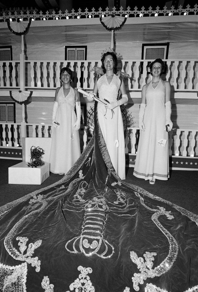 Three women in elegant gowns and gloves stand on a stage. The central figure wears a long cape with a detailed embroidered design, while the other two hold small bouquets. The backdrop resembles a decorative ship deck.